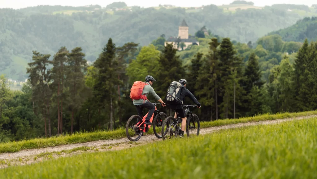 2 Mountainbiker fahren auf einem Waldweg. In der Ferne sieht man ein Schloss aus dem Wald ragen.